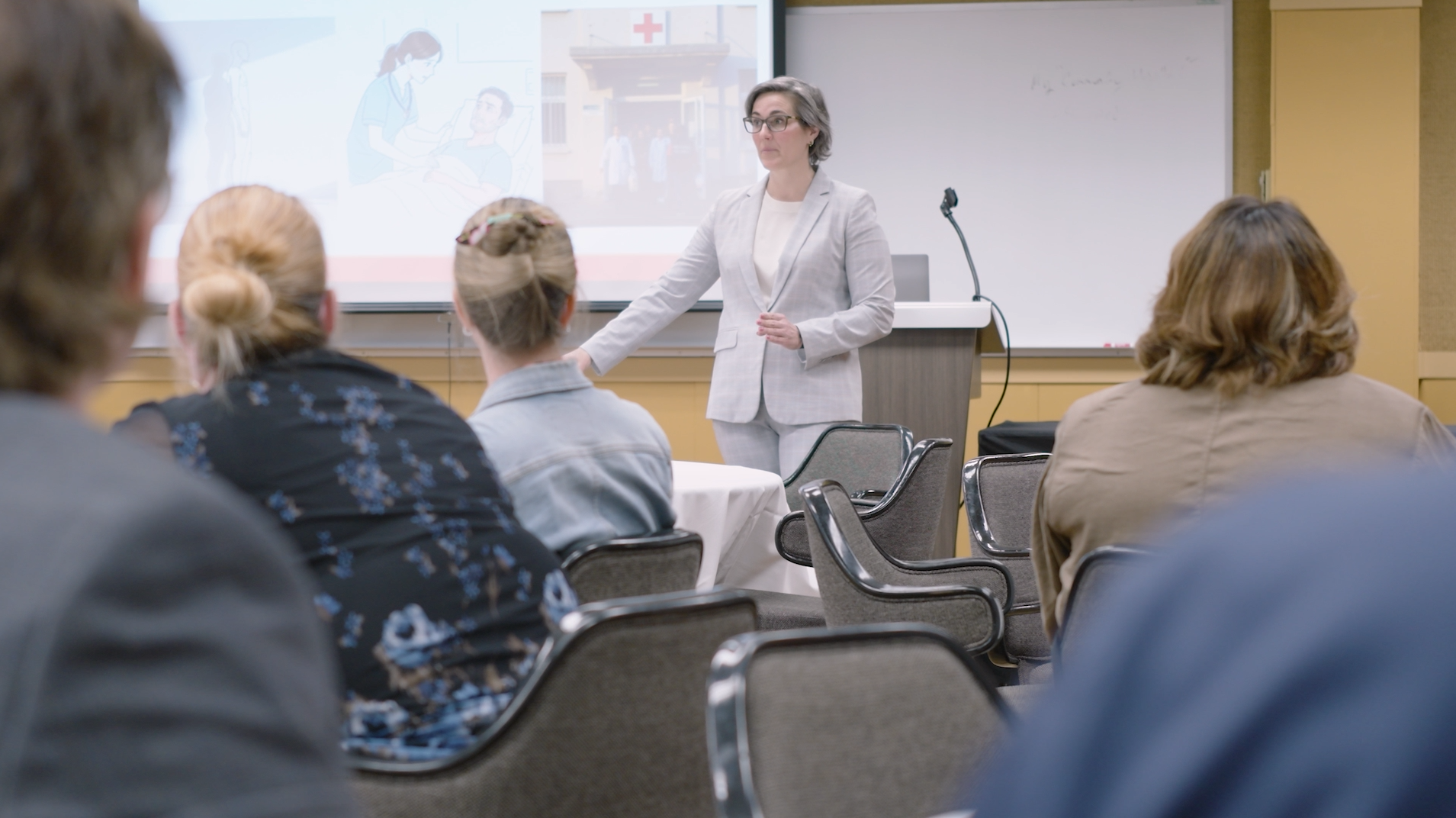 A presenter in a light gray suit stands at the front of a meeting room, speaking to attendees seated at round tables. Behind the presenter, a projected slide displays healthcare-related images, including a patient receiving care and a hospital entrance. A podium with a microphone and a whiteboard are positioned beside the presentation screen, creating a professional educational setting.
